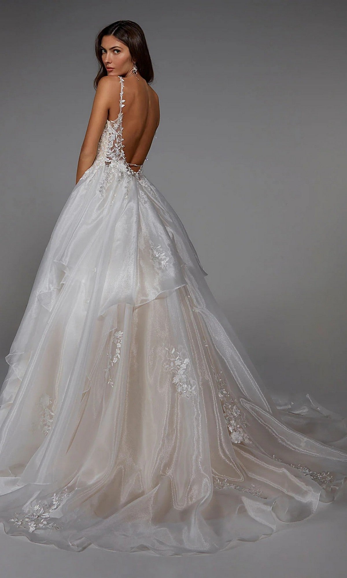 A woman wears the Alyce 7047 long white dress—backless with lace accents, featuring a layered train—against a plain gray background.